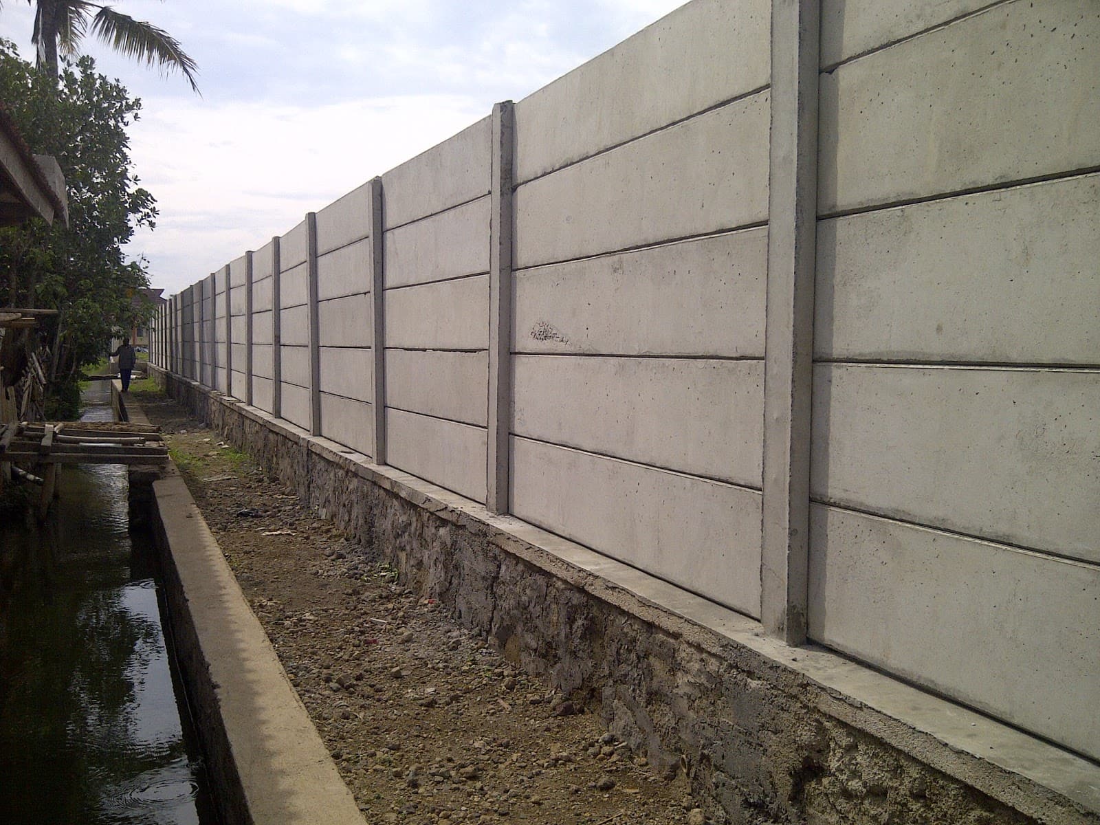 Pagar panel beton precast panjang yang berdiri di atas fondasi batu di samping saluran air.
