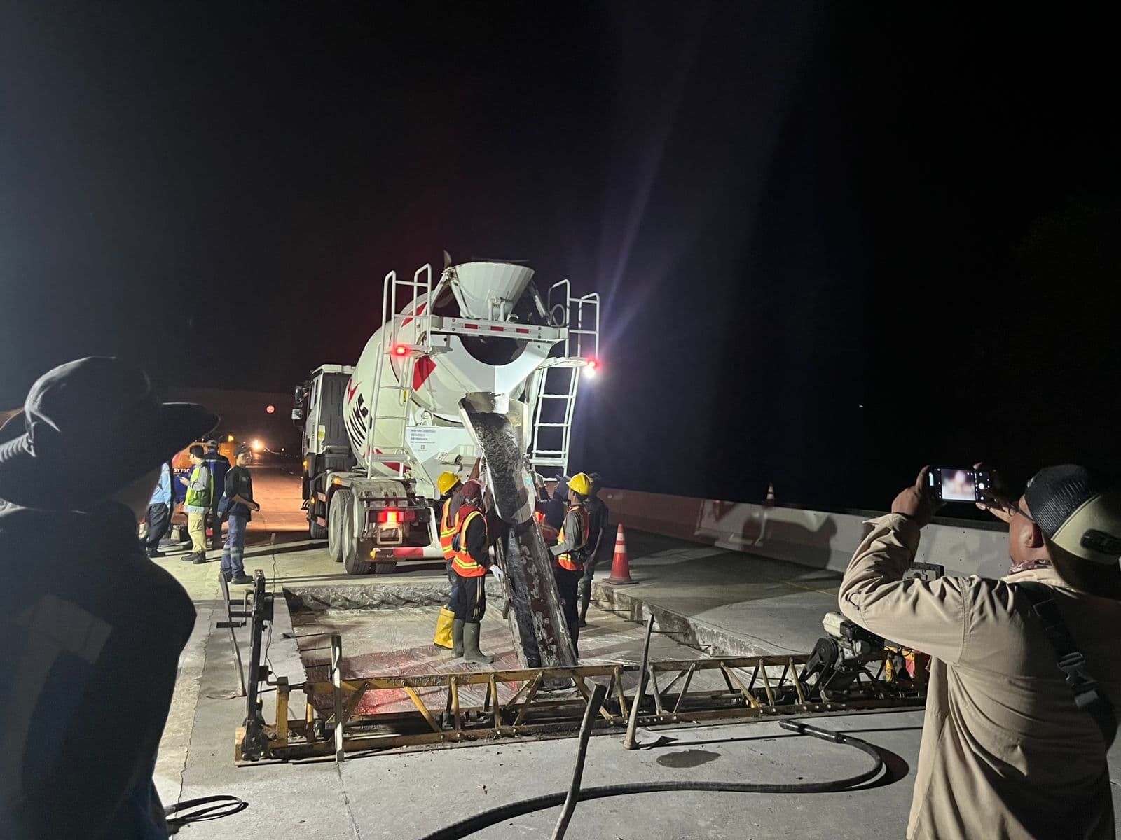 Pekerja konstruksi menuangkan beton dari truk pengaduk ke dalam cetakan jalan pada malam hari.