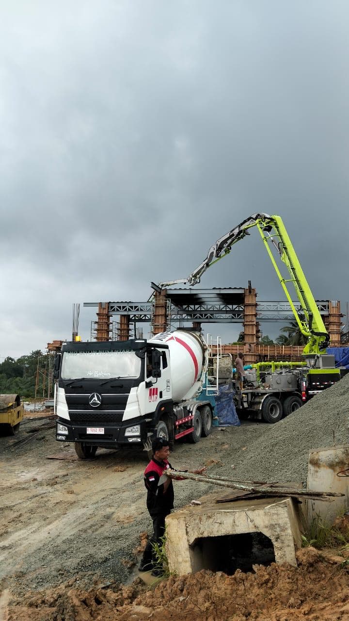 Truk molen putih dan pompa beton hijau bekerja di lokasi pembangunan struktur jembatan layang.