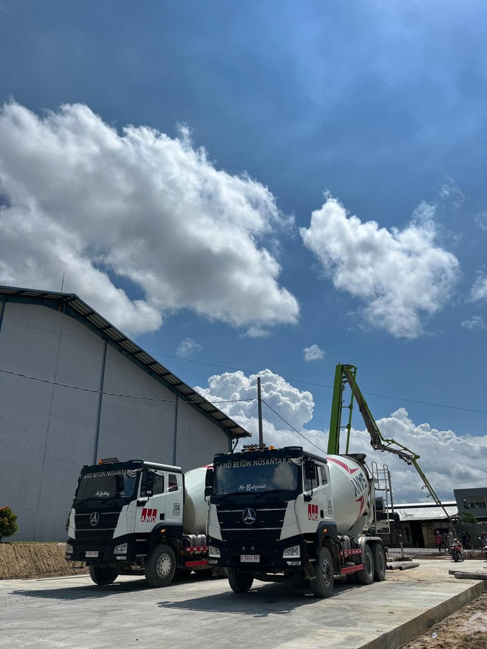 Dua truk pengaduk beton AMD Beton Nusantara parkir di depan gudang saat cuaca cerah.
